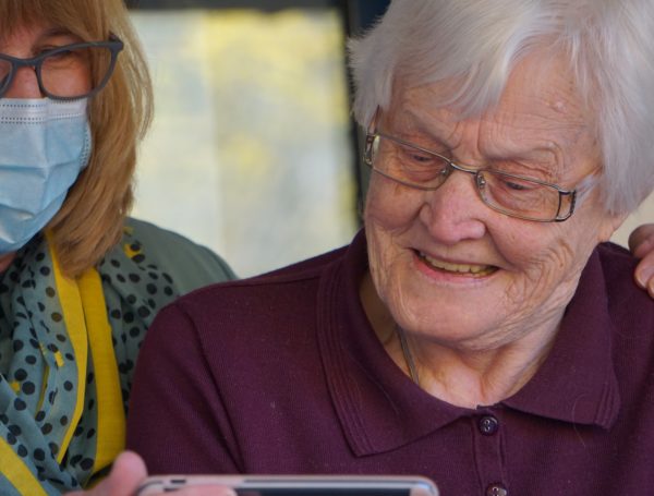 Woman with mask helping elder woman with videocall